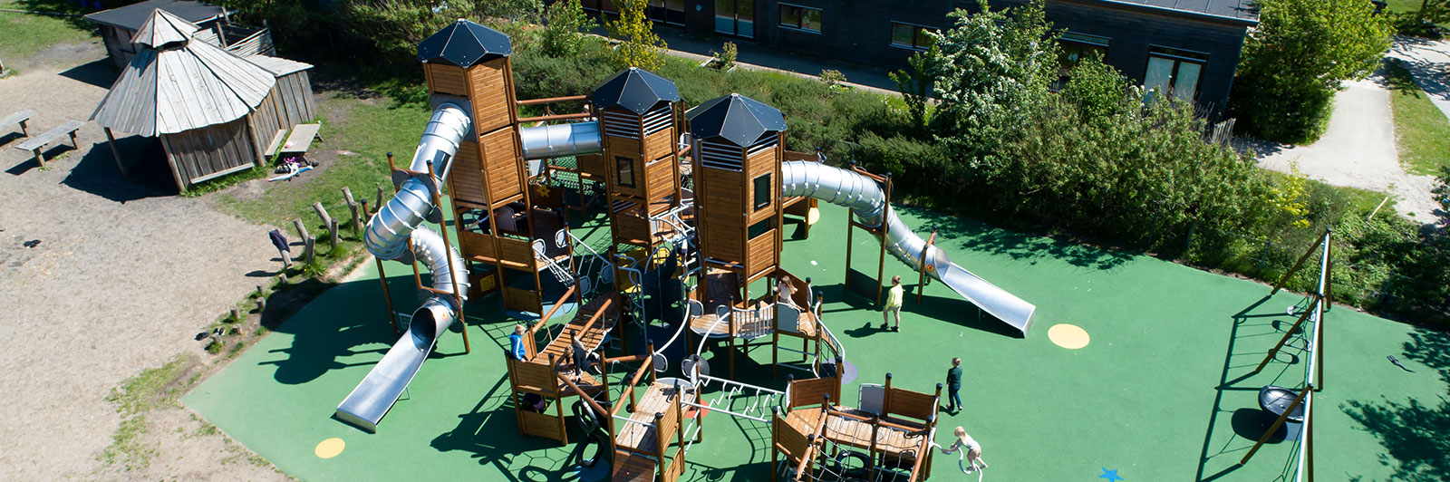 Arial shot of a very large wooden playground tower, it has tube slides, obstacle course sections and many more play opportunities.